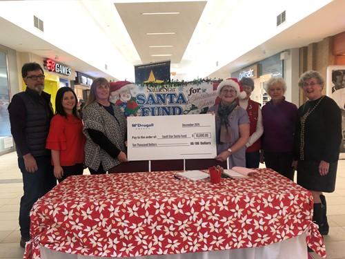 Shows 7 people standing in a shopping mall holding a large cheque from McDougall Energy during Christmas