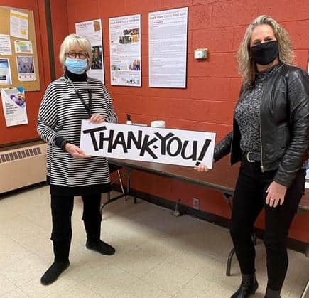 Shows 2 women standing indoors with face masks holding a sign that says Thank You